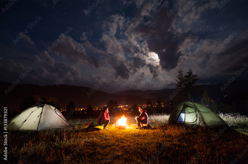 Man and woman sitting at a campfire near two tents in the night under incredibly beautiful a cloudy sky in the background mountains and luminous town