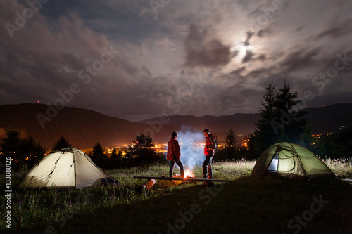 Girl and guy stands near camp and looks on the campfire with smoke under evening cloudy sky on the background mountains and luminous town
