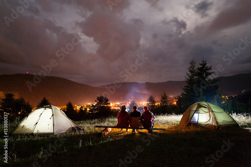 Three tourists sitting on boards around the campfire near tents at night in the background of cloudy sky, mountains and luminous town