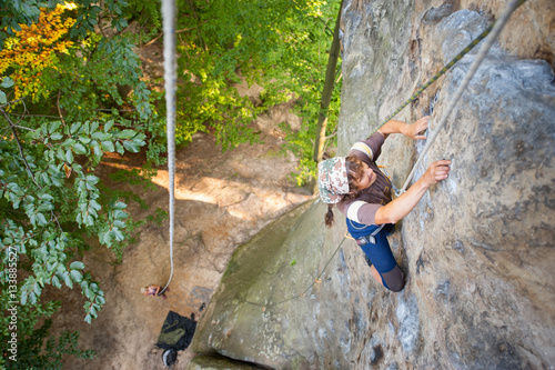 Fototapeta Naklejka Na Ścianę i Meble -  Older female rock climber is climbing with rope on a rocky wall of large boulder. Belayer standing on the ground insuring the climber. View from the top. Extreme sports concept