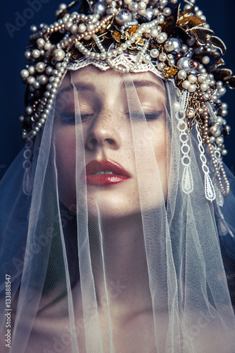 Принти на полотні fashion beauty portrait of young beautiful young woman with makeup and freckles on her face and pearl headpiece on her head and white tulle in front of her face on dark blue background