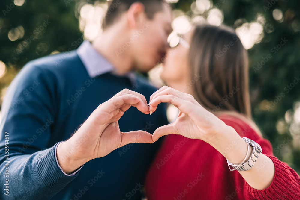 Young, happy couple enjoying outdoors. They making heart shape with hands and kissing. Selective focus on hands.