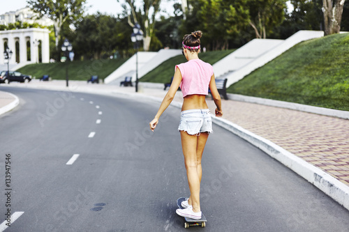 Beautiful hipster girl in a jeans shorts and pink shirt walking with skateboard near the green park  sexy style  adult  long legs  brunette  white shoes  sexy model  posing on camera