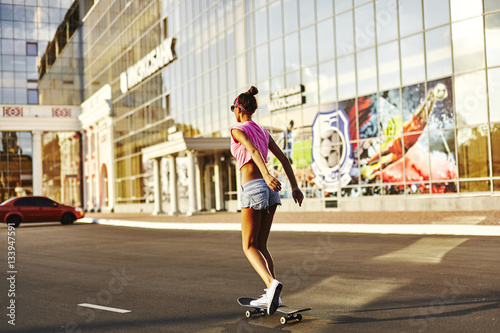 Beautiful hipster girl in a jeans shorts and pink shirt walking with skateboard near the green park, sexy style, adult, long legs, brunette, white shoes, sexy model, posing on camera © evgeniy_regulyan