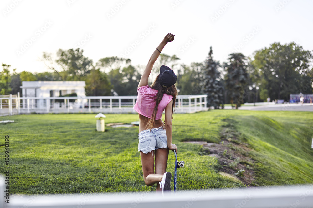 Beautiful hipster girl in a jeans shorts and pink shirt walking with skateboard near the green park, sexy style, adult, long legs, brunette, white shoes, sexy model,  posing on camera