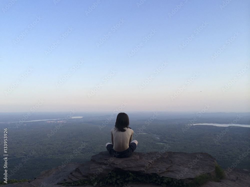 Rear view of shirtless boy sitting on cliff against clear blue sky