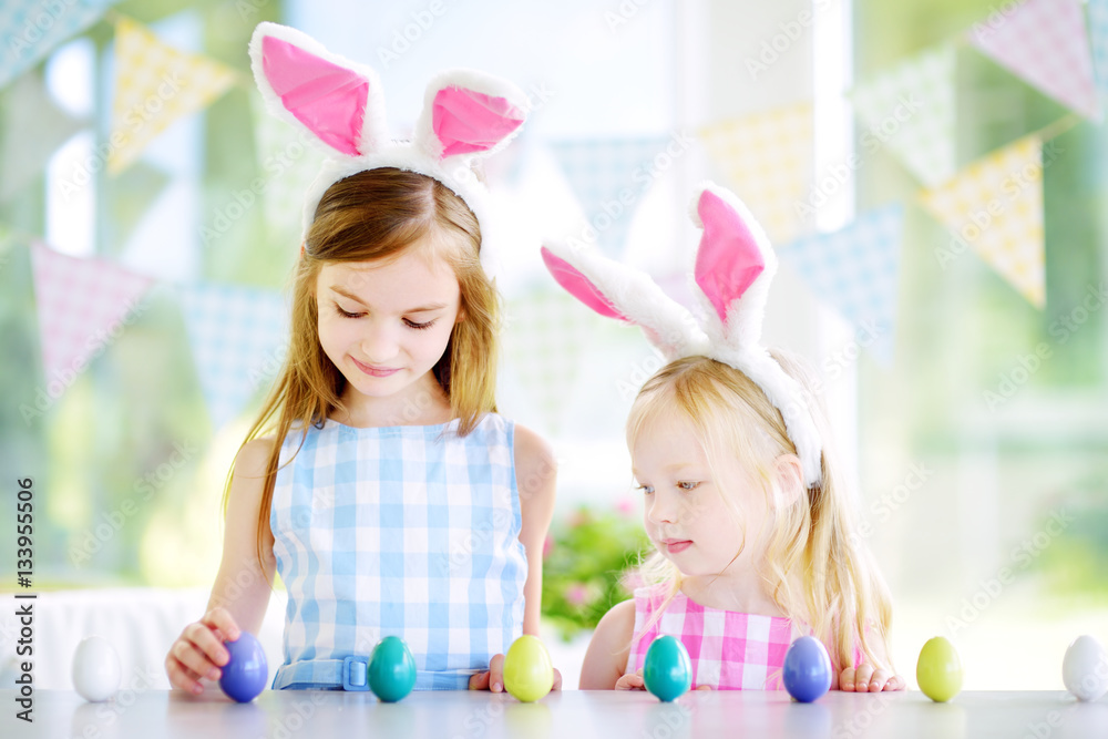 Two cute little sisters wearing bunny ears playing egg hunt on Easter Two cute little sisters wearing bunny ears playing egg hunt on Easter