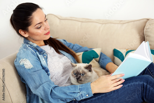 Young woman reading book while sitting on sofa with cute cat