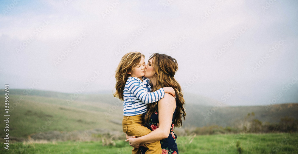 Loving woman kissing daughter while standing on field against sky Loving woman kissing daughter while standing on field against sky