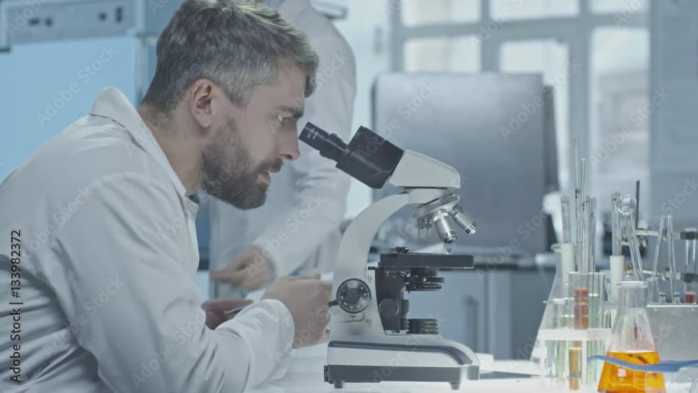 Male scientist with beard looking into microscope, then writing down notes in clipboard handed by female assistant Male scientist with beard looking into microscope, then writing down notes in clipboard handed by female assistant