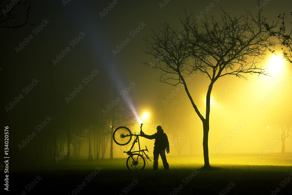 Naklejka premium One man is standing and holding the bicycle in foggy and mysterious park at night