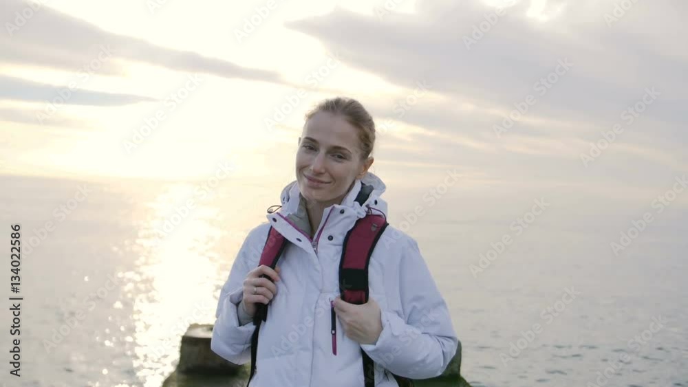 Portrait of beautiful young smiling woman in jacket with backpack on pier with sea on a background Portrait of beautiful young smiling woman in jacket with backpack on pier with sea on a background