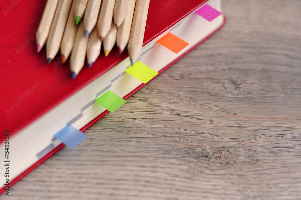 Obraz premium Red diary notebook and colored pencils on the desk wood texture.