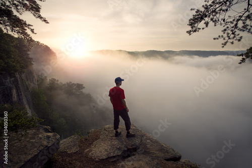 A white man wearing a red shirt looks out over the New River Gorge in West Virginia on a misty morning at sunrise.