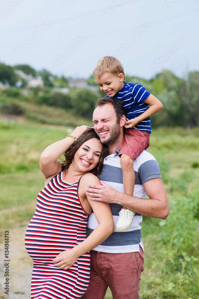 Pregnant family of three in expectation of baby. Father mother and son playing together in day outside. Happy people hugging. Smiling laughing baby sitting on shoulders of daddy. Pregnant family of three in expectation of baby. Father mother and son playing together in day outside. Happy people hugging. Smiling laughing baby sitting on shoulders of daddy.