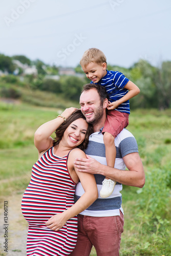 Pregnant family of three in expectation of baby. Father mother and son playing together in day outside. Happy people hugging. Smiling laughing baby sitting on shoulders of daddy.