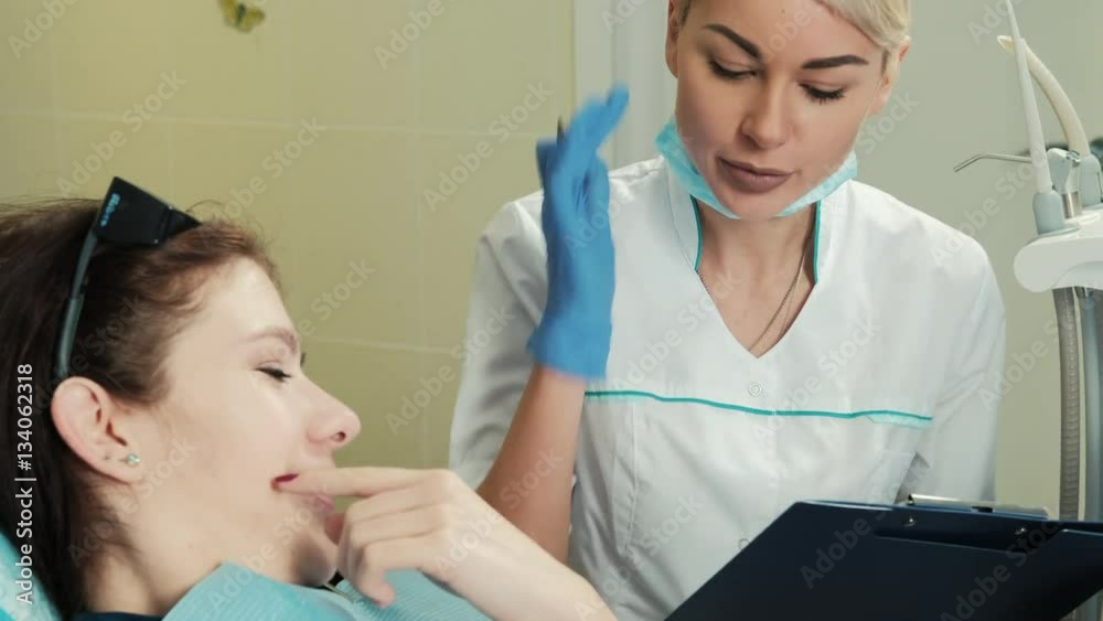 Medic polls your patient about the history of dental disease. Woman with dark hair sitting in a dental chair and responds to medical questions that she asks with a notebook and pen in hand. The doctor Medic polls your patient about the history of dental disease. Woman with dark hair sitting in a dental chair and responds to medical questions that she asks with a notebook and pen in hand. The doctor