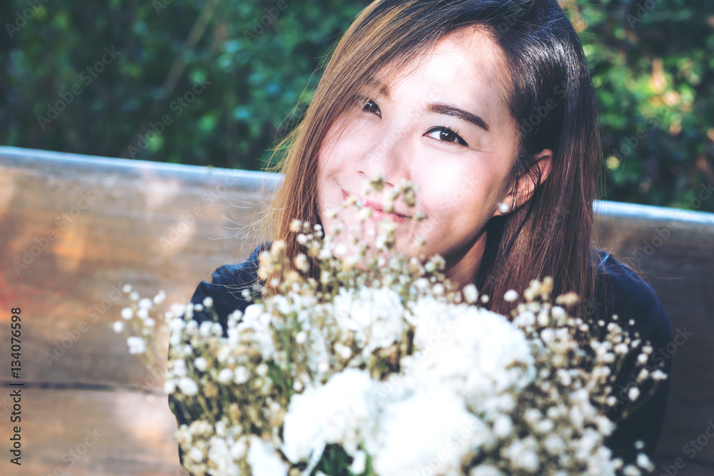 A beautiful asian woman and white flowers with feeling happy and smiley face , sitting on wooden chair in green nature outdoor background A beautiful asian woman and white flowers with feeling happy and smiley face , sitting on wooden chair in green nature outdoor background