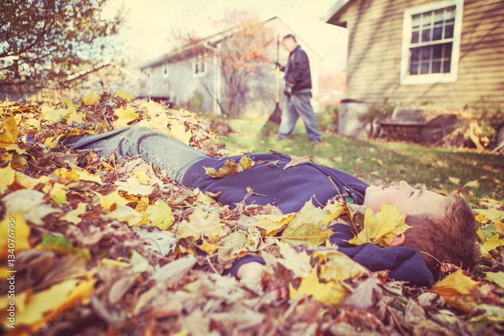 Fototapeta premium Young boy daydreaming in a pile of fall leaves while his father
