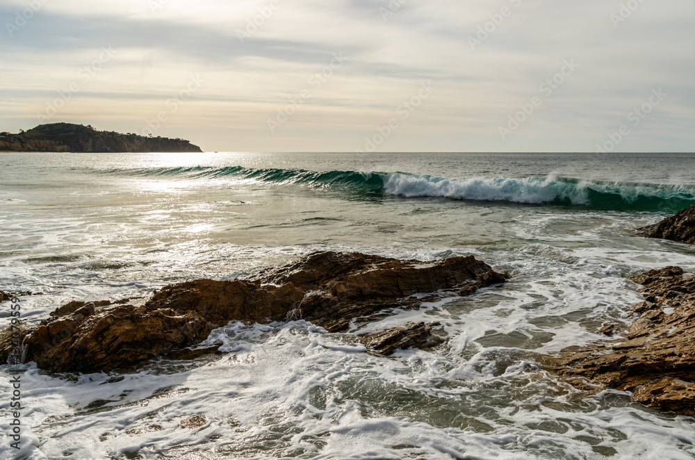 Waves of the calm Pacific Ocean gently break just beyond a rocky section of shoreline at Crystal Cove State Park in Laguna Beach, California.
