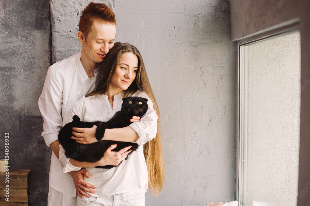 Happy young beautiful caucasian couple hugging and holding a cat. Playing with pet at home. Love, relationships, coziness, leisure, animal protection concept.