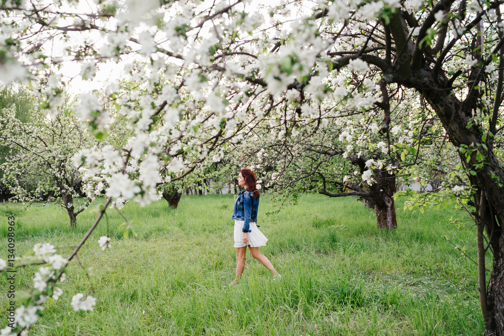 Young pretty caucasian woman in white dress and denim jacket walks in a blooming spring garden on sunset. Youth, freshness, beauty, romance, happiness concept.