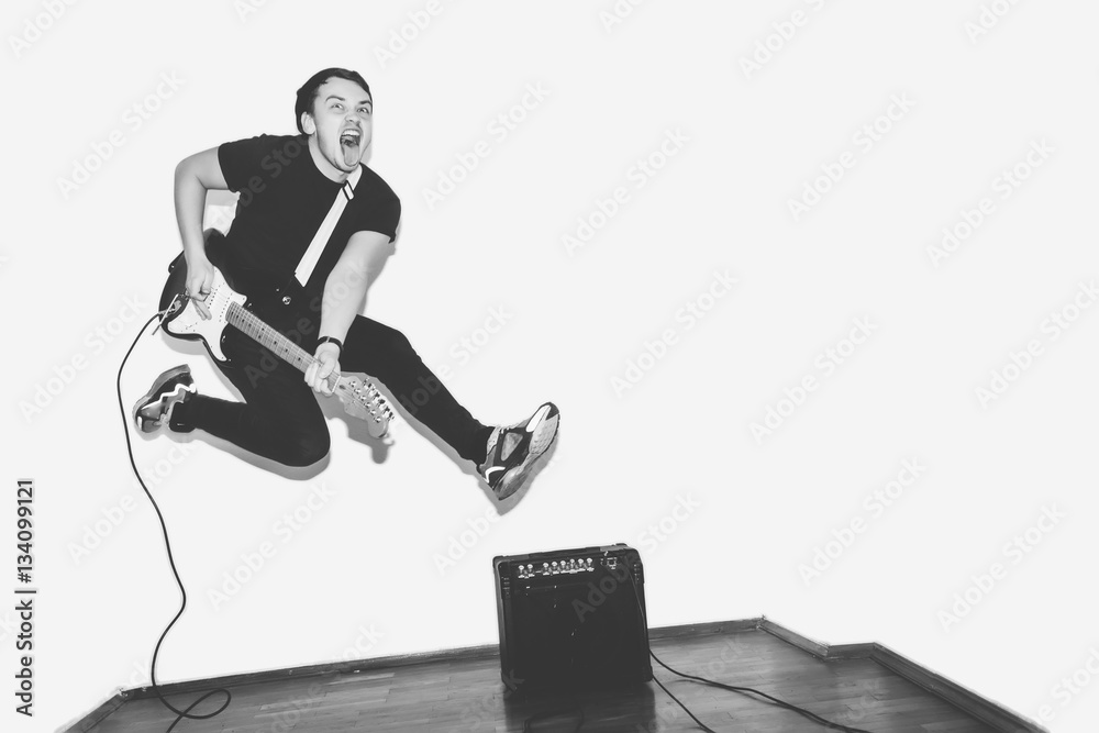 Fototapeta premium Awesome crazy fashion young musician rock guitar player jumps with passion in studio. Stylish rocky emotional man. Black and white toned.