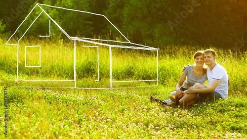 Young happy couple sitting on green grass in park and dreaming about they own house. Mortgage concept.