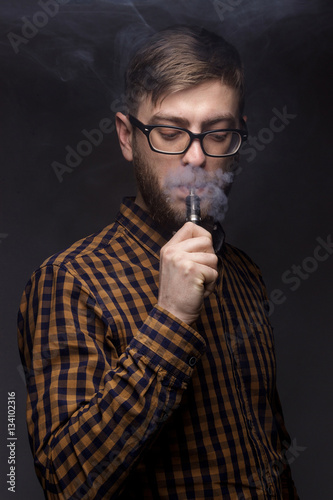 man with a beard wearing glasses on black background smoke Vape. large portrait of . electronic cigarette. E- for vaping. Popular devices the year - modern device. Stop smoking,