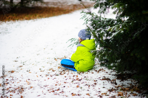 The child in bright ski suit alone sits on snow under a tree.