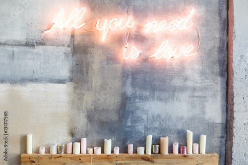 Neon inscription "All you need is love" on a grey concrete wall and many candles under them