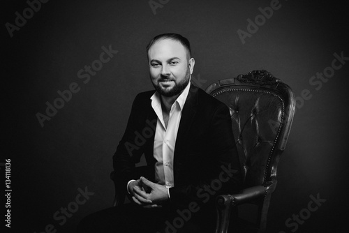 portrait of young man in black suit sitting on luxury chair on dark background monochrome © k8most