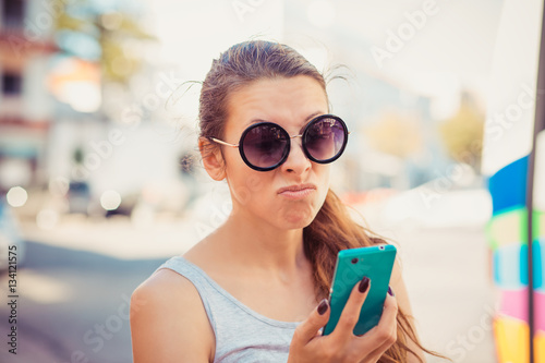 Portrait of an annoyed and frustrated young woman on the phone isolated on city scape background. Human emotion face expression feeling
