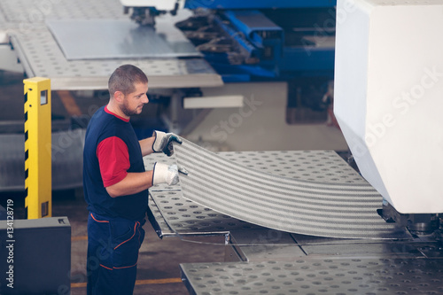 Inside a factory, industrial worker in action on metal press machine holding a piece of steel ready to be worked.