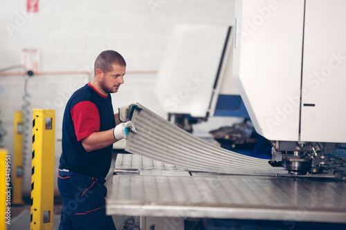 Fotografie Inside a factory, industrial worker in action on metal press machine holding a piece of steel ready to be worked