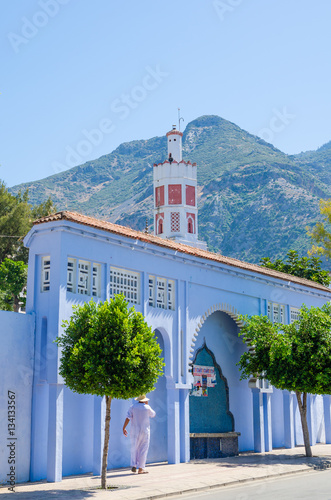 Arabic man standing in front of blue washed wall, red mosque and mountain in the town Chefchaouen, Morocco © Fabian