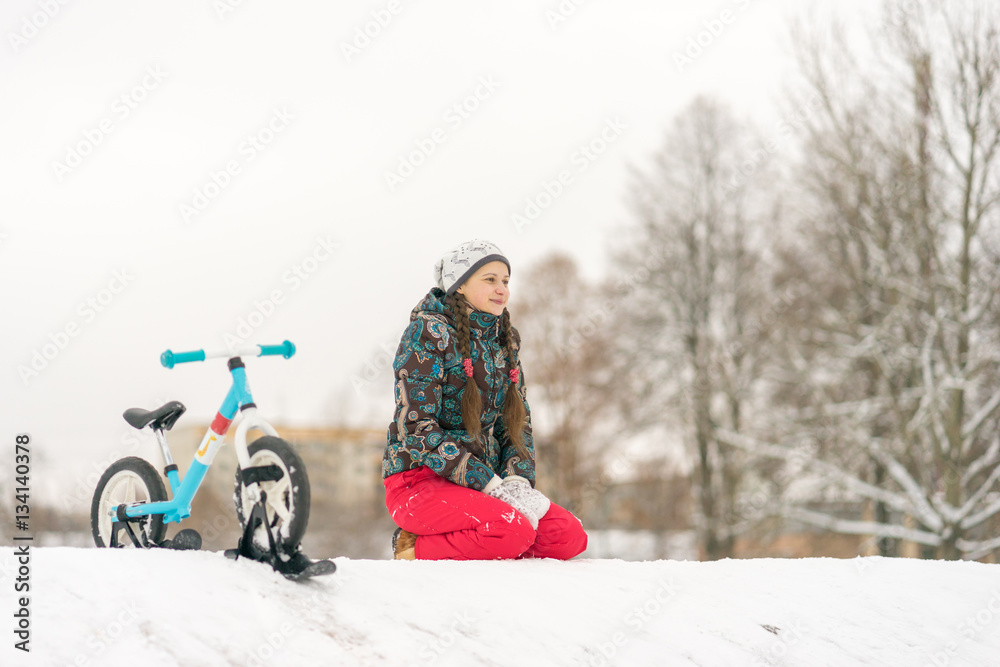 Young girl sitting on a snowy mountain near the bike with skis o