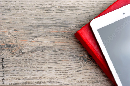 Red notebook and white tablet on a table with a wooden texture.