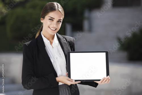 Beautiful young business woman is holding a tablet pc, with a blank screen, horizontally, while smiling photo