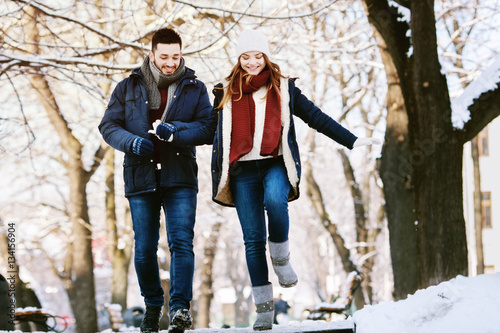 Outdoor full body portrait of young happy beautiful smiling couple walking in park. Models wearing stylish warm winter clothes. Day light, sunny weather. Trees with snow on background