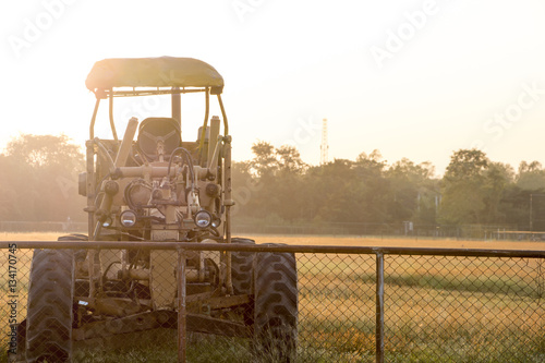 An old tractor or grader machine parked behind a metallic mesh fence with back-lit, glowing setting sun