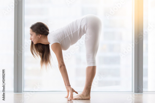 Young woman practicing yoga, standing in head to knees, Ardha uttanasana exercise, Half forward bend pose, working out, wearing sportswear, white t-shirt, pants, indoor full length, near floor window