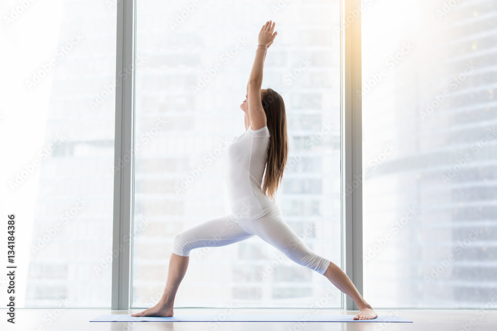 Young attractive woman practicing yoga, standing in Warrior one exercise, Virabhadrasana I pose, working out, wearing sportswear, white t-shirt, pants, indoor full length, floor window with city view Young attractive woman practicing yoga, standing in Warrior one exercise, Virabhadrasana I pose, working out, wearing sportswear, white t-shirt, pants, indoor full length, floor window with city view