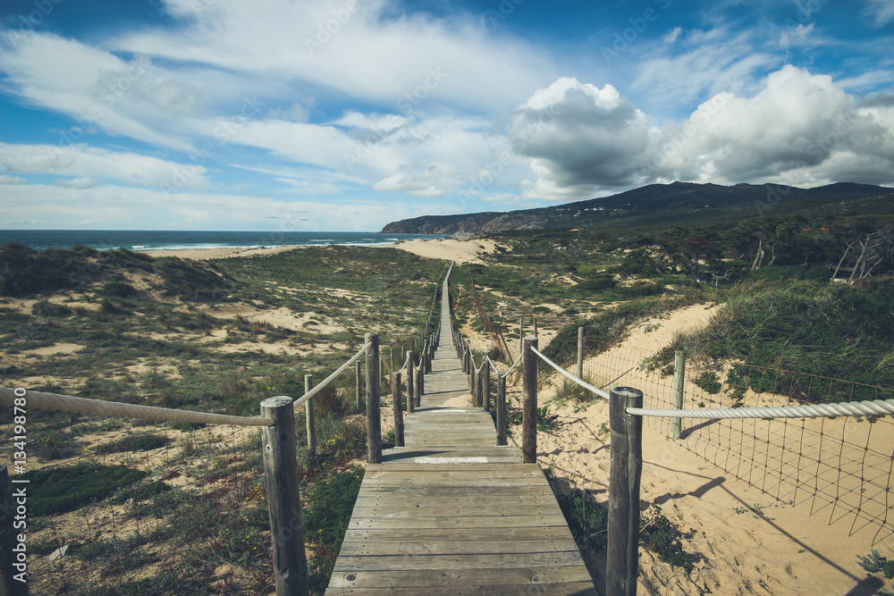 Wooden path to the beach on the dunes with a mountain and the ocean in background. Guincho beach in Cascais, Portugal. Vintage filter