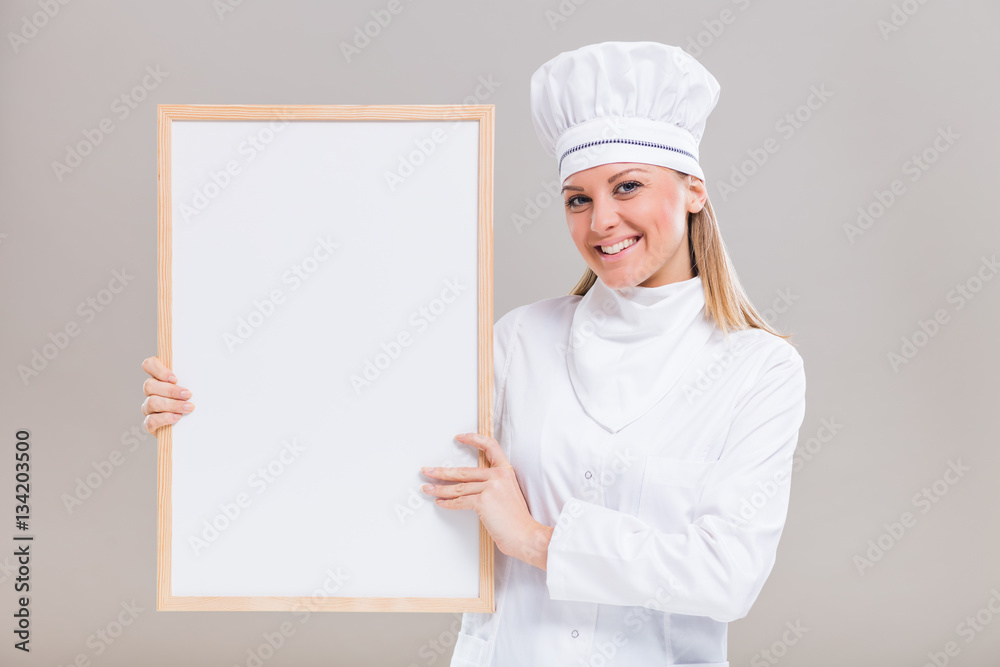 Portrait of beautiful female chef holding whiteboard on gray background.
Portrait of beautiful female chef holding whiteboard on gray background.