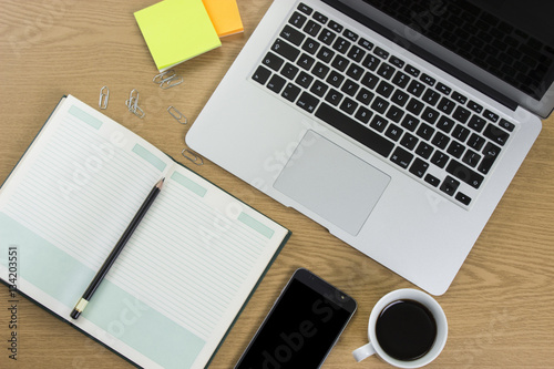 Modern wooden office desk table with laptop, smartphone and other supplies with cup of coffee. Blank notebook page for input the text in the middle. Top view, flat lay.