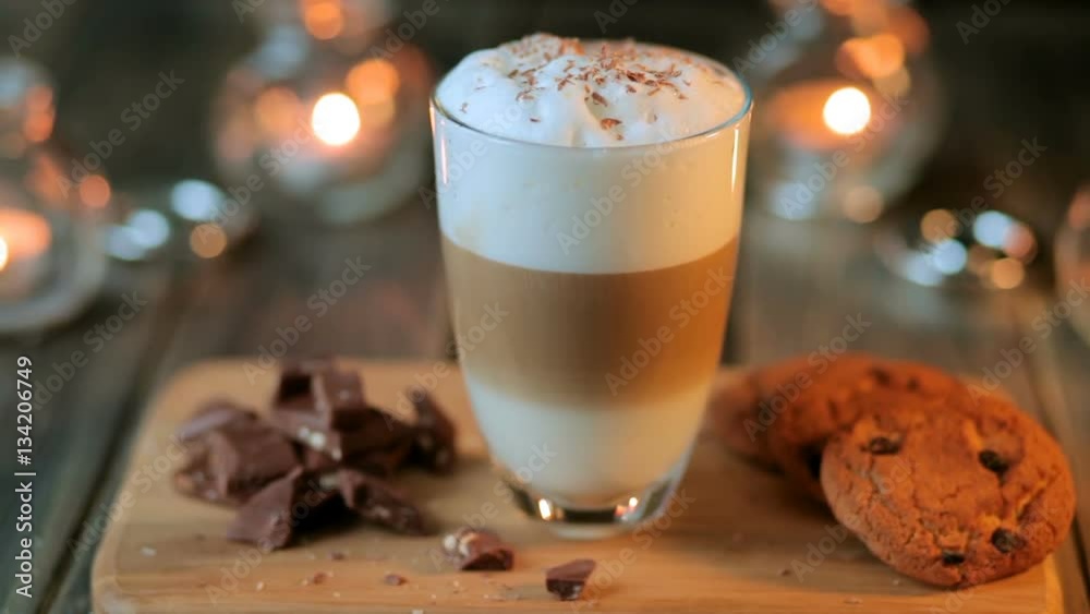 Beautiful composition - hot coffe latte cappucino with oatmeal cookies and chocolate pieces in a transparent glass. The glass stands on a wooden stand. Behind the candles are burning Beautiful composition - hot coffe latte cappucino with oatmeal cookies and chocolate pieces in a transparent glass. The glass stands on a wooden stand. Behind the candles are burning