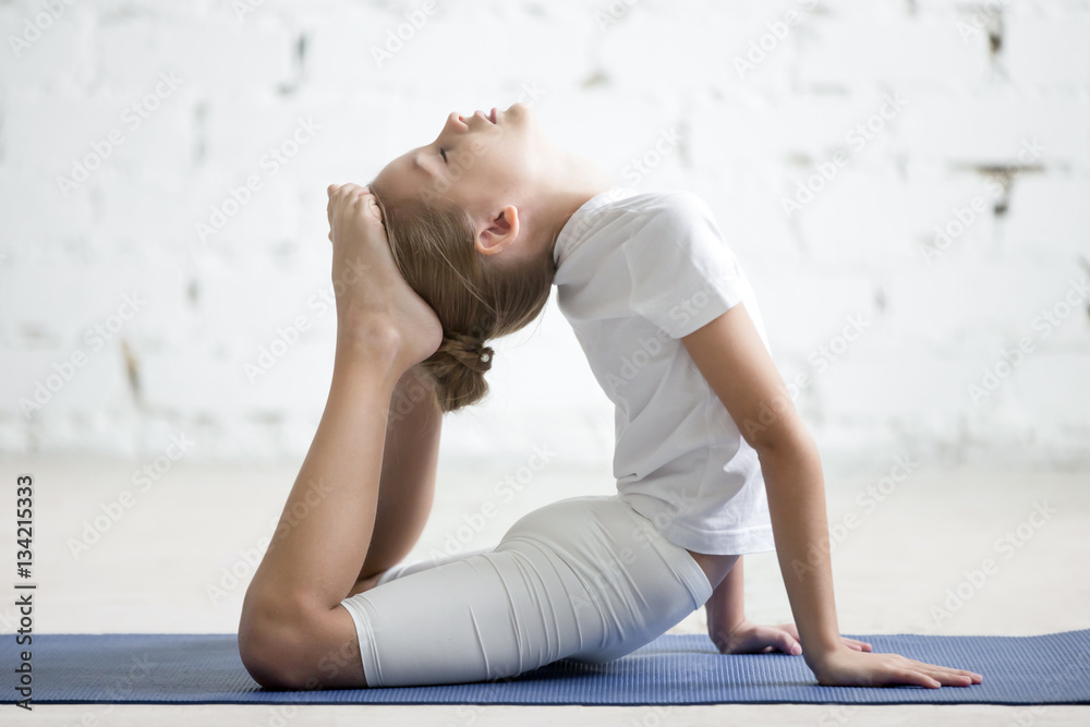 Fototapeta premium Girl child practicing yoga on blue mat, stretching in raja bhudjangasana exercise, King Cobra pose, working out wearing sportswear, t-shirt, pants, indoor full length, white loft studio background