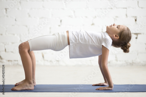Serious girl child practicing yoga, standing in Reverse Table Top exercise, Bridge pose working out wearing sportswear, t-shirt, pants, indoor full length, white loft studio background