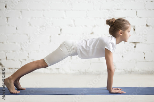 Side view of girl child practicing yoga, standing in phalankasana pose, Push ups or press ups exercise, working out wearing sportswear, t-shirt, pants, indoor full length, white loft studio background